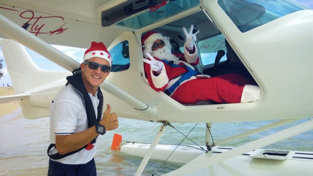 One person in a Santa hat gives a thumbs up outside a seaplane, while Santa makes peace signs inside.